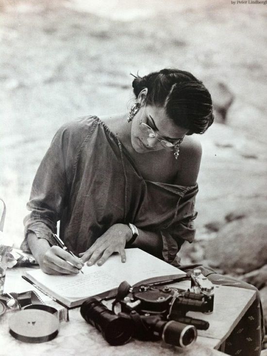 a woman sitting at a table writing in a book with old cameras and other items around her