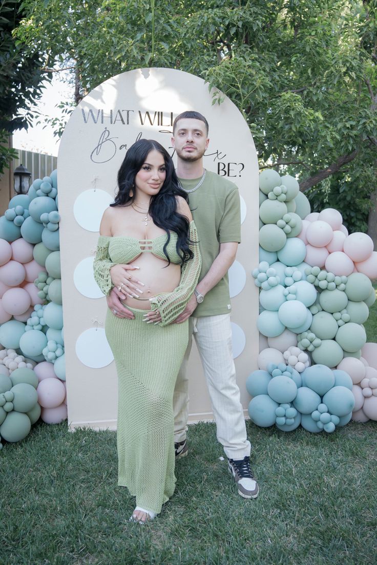 a pregnant couple standing in front of an outdoor backdrop with balloons and flowers on it