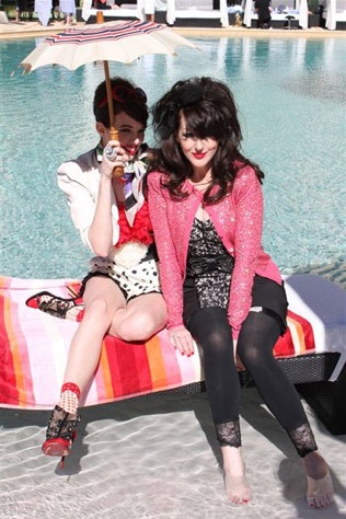 two women sitting on a beach towel next to a pool with an umbrella over them