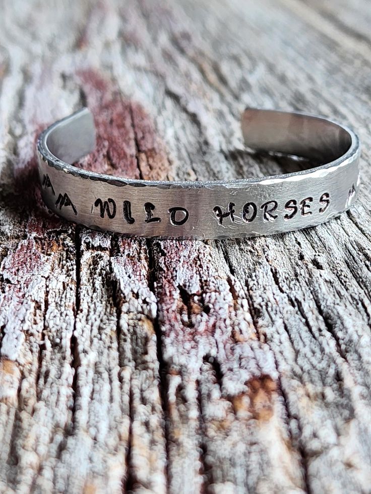 Handstamped "Wild Horses" Stainless steel Cuff Bracelet, handmade with hammered edges and mountains stamped on either side and a sunset on one side .  Pictures of additional bracelets are sold separately.  This Cuff will fit a wrist size 5.75"-7" and will come packaged in a pretty floral gift bag.  Please Note:  This is a handstamped piece.  It is stamped with irregularities, that's what leads to its perfectly imperfect charm. If you would like a custom cuff bracelet with your message just message me to place your personal order. Stay connected: Give me a "Follow" and "like" for Specials and updates! Instagram: https://www.instagram.com/strandsbyjm Facebook: https://www.facebook.com/strandsbyjm Thank you for Shopping Strands by Jm! Jewelry Care Recommendations.  Apply cosmetics, hair produ Horses And Mountains, Mountain Stamp, Metal Stamped Bracelet, Cowgirl Bracelets, Custom Cuff Bracelet, Jewelry Rustic, Stamped Bracelet, Floral Gifts, Stay Connected