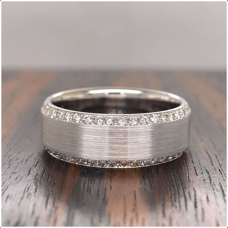 a white gold wedding band with diamonds on it sitting on a wooden table in front of a gray wall