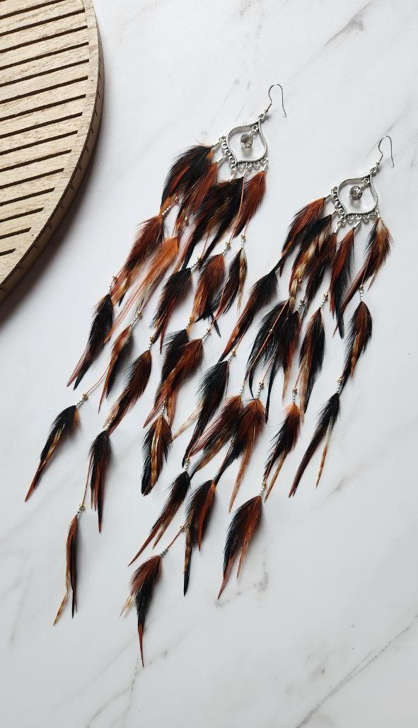 Unleash your wild side with these stunning handmade earrings featuring an array of vibrant brown and black feathers. These long dangle earrings are adorned with delicate beads and metallic accents, adding a touch of tribal flair to their bohemian elegance. Perfect for festival outfits, boho-chic style, or as a unique gift, these lightweight earrings create a dramatic and eye-catching effect. Embrace the spirit of adventure and make a bold statement with these one-of-a-kind feather earrings! Add Brown Feather Dangle Earrings, Bohemian Black Feather Earrings, Feather Extensions, Boho Feathers, Free Spirit Style, Long Dangle Earrings, Lightweight Earrings, Black Feathers, Feather Earrings