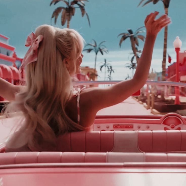 a woman sitting in the back of a pink car with palm trees and buildings behind her