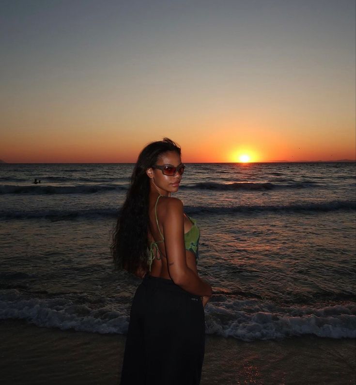 a woman standing on top of a beach next to the ocean at sun set with her hair blowing in the wind