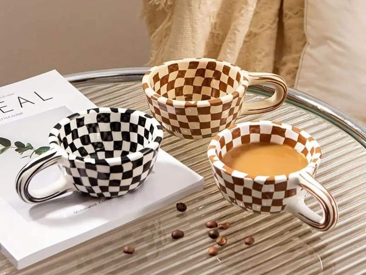 two coffee cups sitting on top of a glass table next to a book and some beans