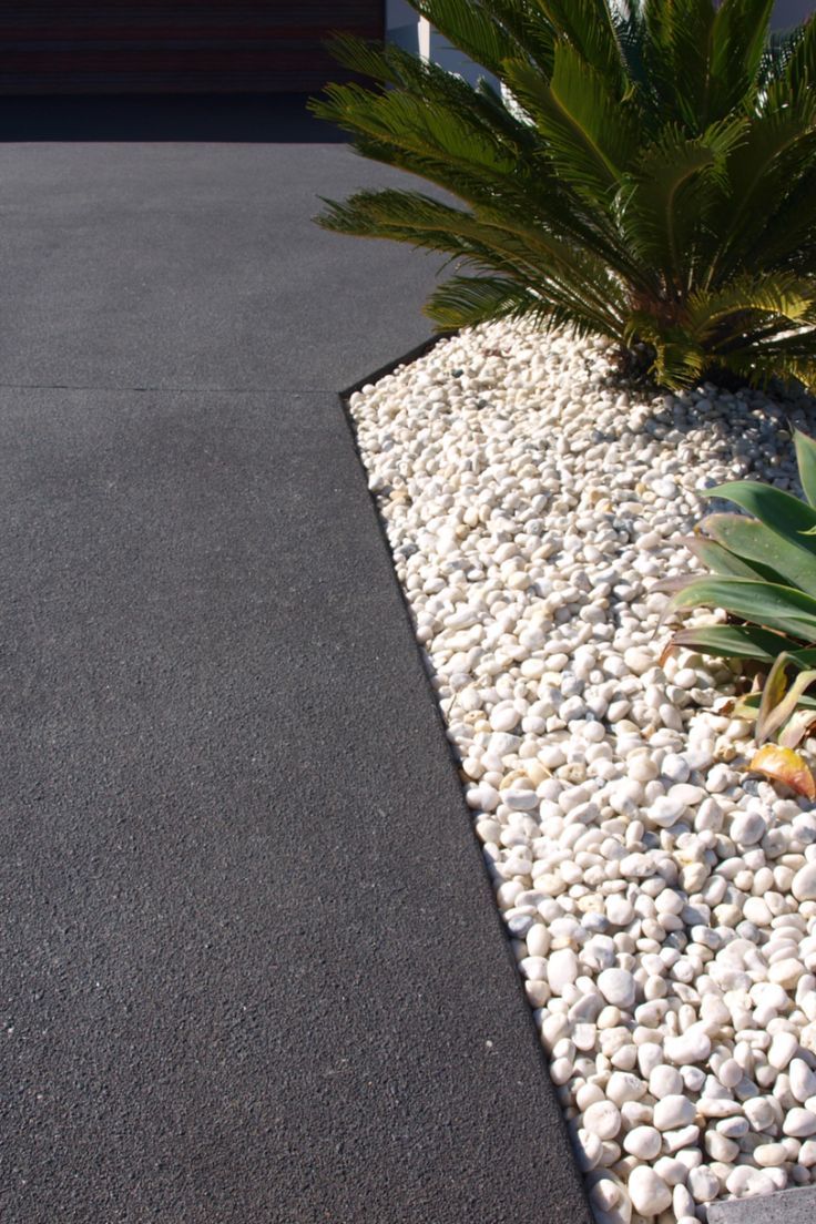 This image shows a concrete driveway coloured Jet Black with a Dulux Avista Colour Hardener. The dramatic tone creates a stunning contrast against the white pebbles in the garden bed. Pebble Driveway, Colored Concrete Patio, Driveway Paint, Cement Driveway, Front Garden Ideas Driveway, Poured Concrete Patio, Concrete Pathway, Concrete Backyard, Modern Driveway