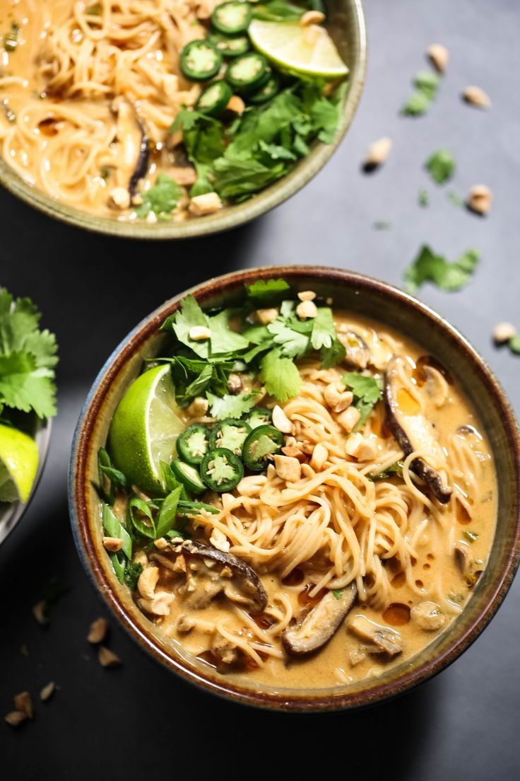 two bowls filled with noodles and vegetables on top of a black table next to limes