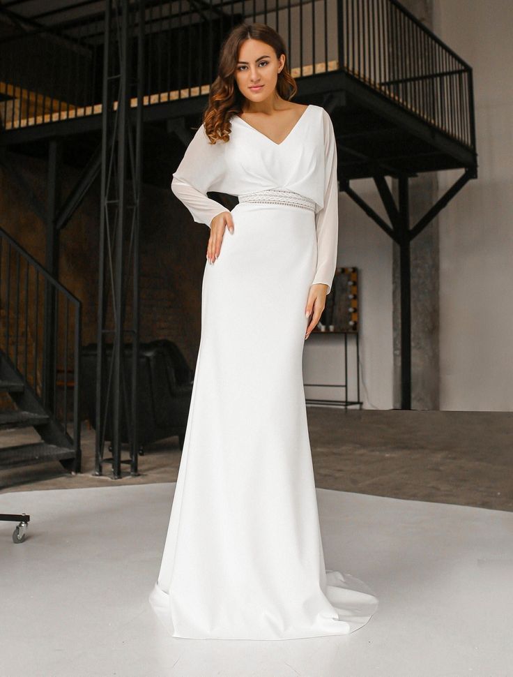 a woman standing in front of a staircase wearing a white dress with long sleeves and belt