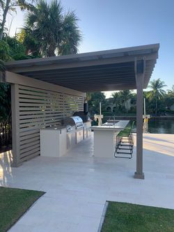 an outdoor kitchen with grills and chairs under a pergolated roof next to a body of water