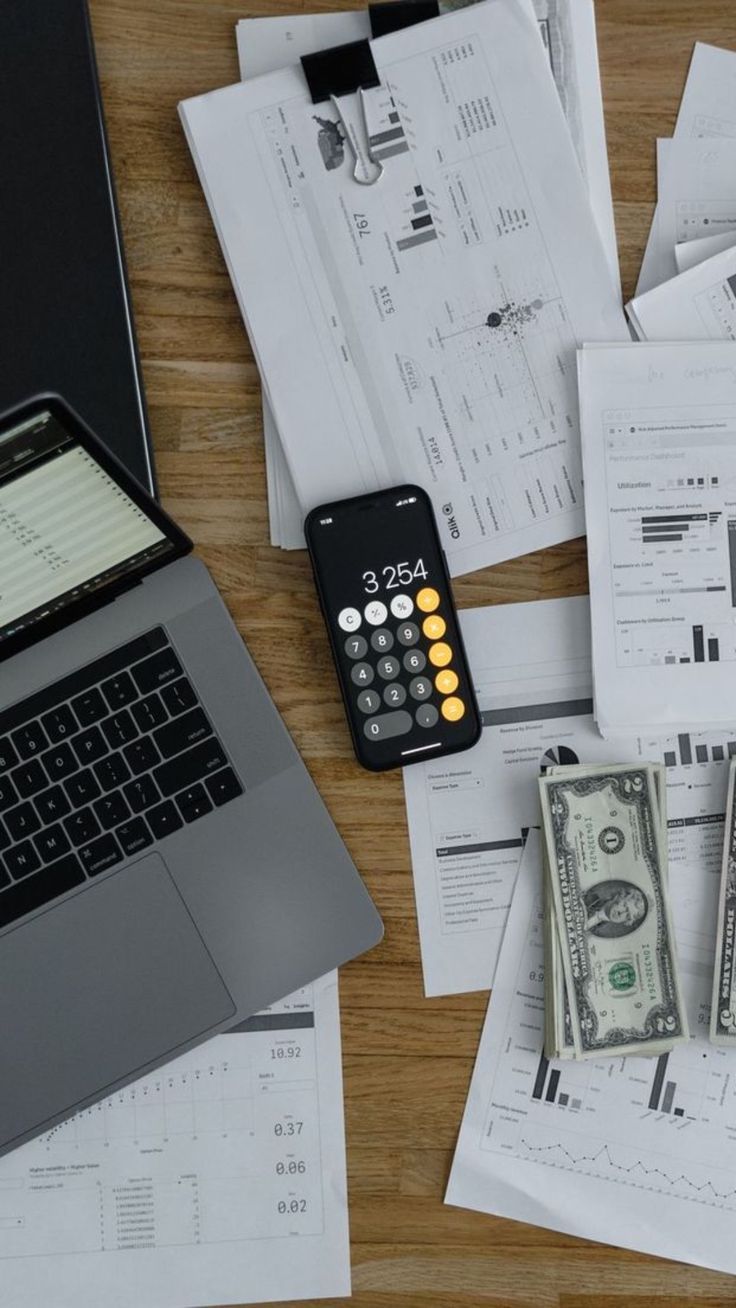 an open laptop computer sitting on top of a wooden table covered in papers and money