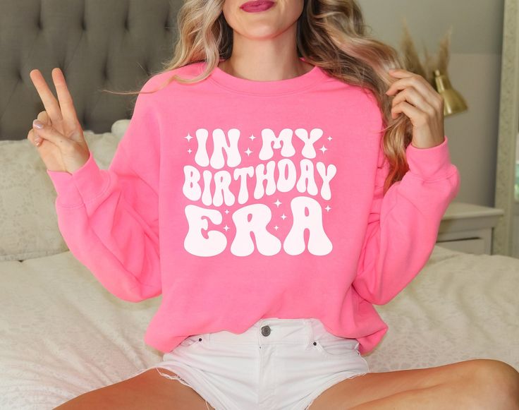 a woman sitting on top of a bed wearing a pink shirt with the words in my birthday era written on it