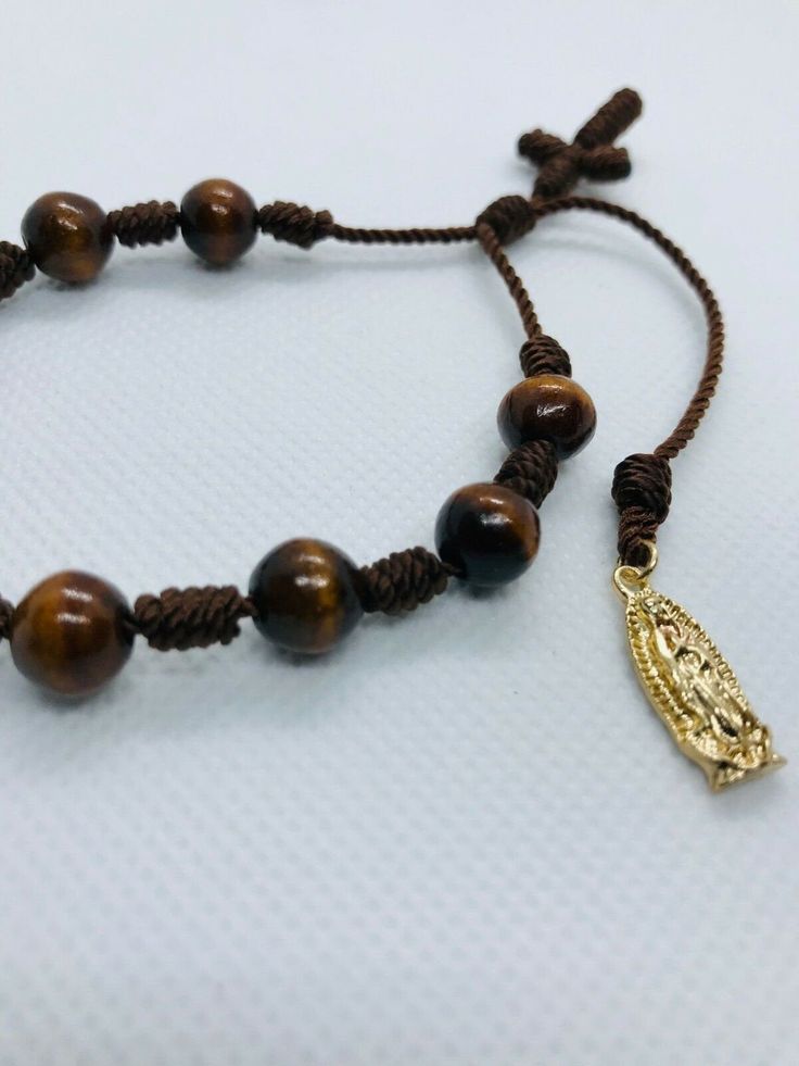 "Handmade brown bracelet with wood beads as denarius 8mm, 10 knots with string #18 Adjustable Virgin of Guadalupe bracelet, plated gold pendant 1.8cm and crucifix made with knots. Bracelet length: from 7¨inches up to 8.5¨inches. Ajustable pulsera de color cafe con Denario de bolas de madera barnizado de 8mm hecha a mano con nudos en hilo cafe reforzado #18 Pulsera religiosa con una medalla de oro laminado de la Virgen de Guadalupe 1.8 cm y un crucifico de nudos. Medida de la Pulsera con minimo c Spiritual Brown Rosary Bracelet With Beads, Spiritual Brown Beaded Bracelets With Wooden Beads, Spiritual Brown Bracelets With Wooden Beads, Spiritual Brown Wooden Beaded Bracelets, Adjustable Rosary Bracelet With Polished Beads, Spiritual Brown Rosary Bracelet With Wooden Beads, Brown Wooden Beads For Bracelets, Brown Wooden Beaded Bracelet, Adjustable Spiritual Rosary Bracelet With Polished Beads