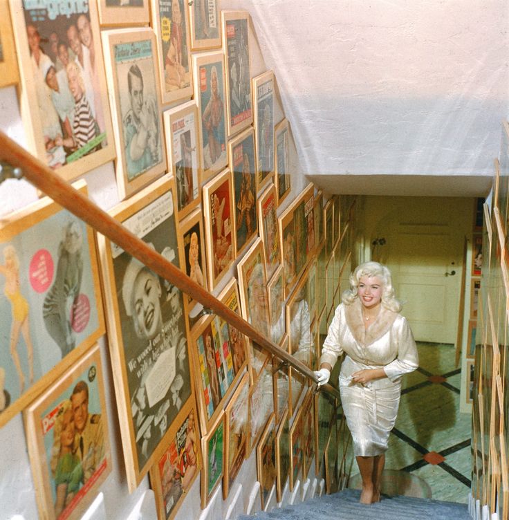 an old woman walking down the stairs in front of many framed pictures and posters on the wall