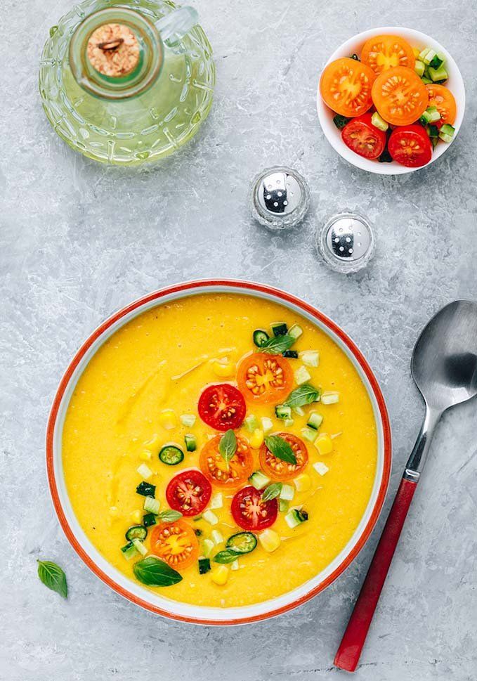 a bowl of soup with tomatoes, corn and basil on the side next to two spoons