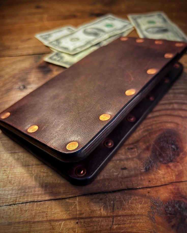 a brown leather wallet sitting on top of a wooden table next to one hundred dollar bills