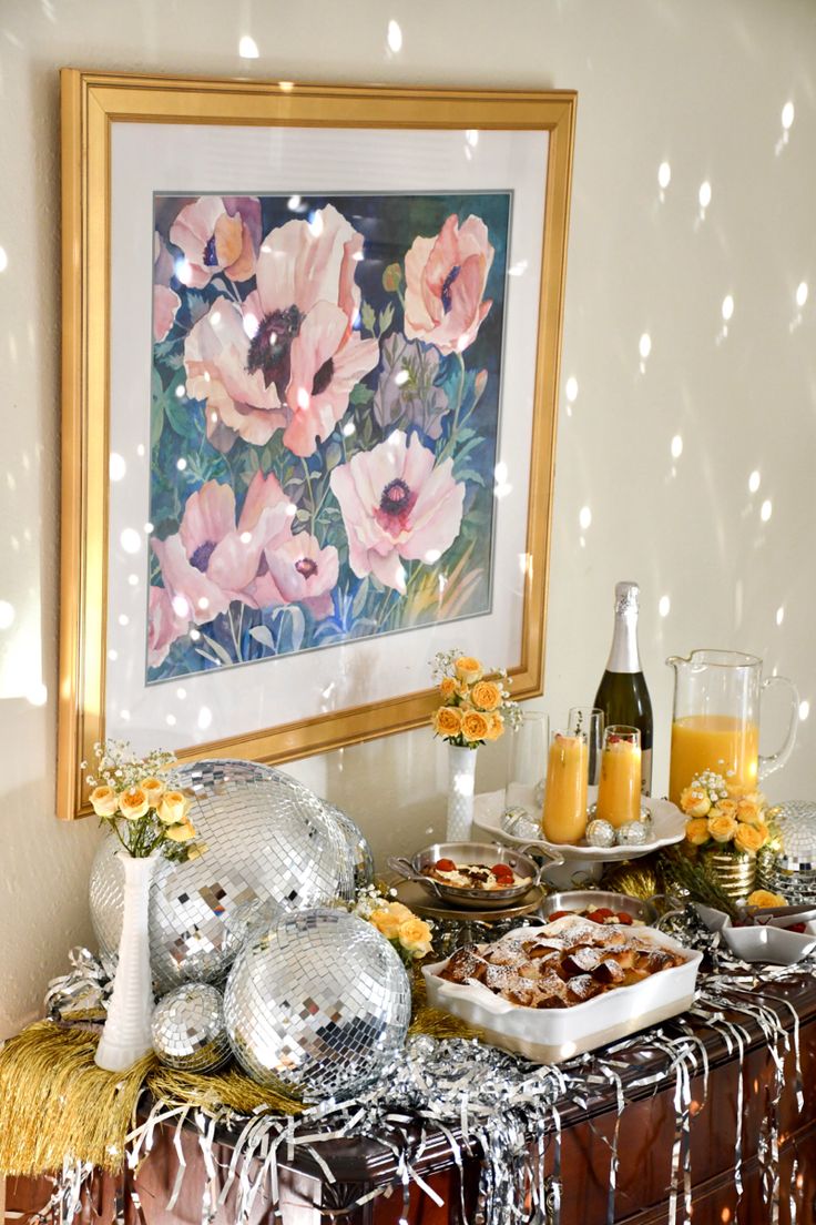 a table with food and drinks on it in front of a framed floral art piece