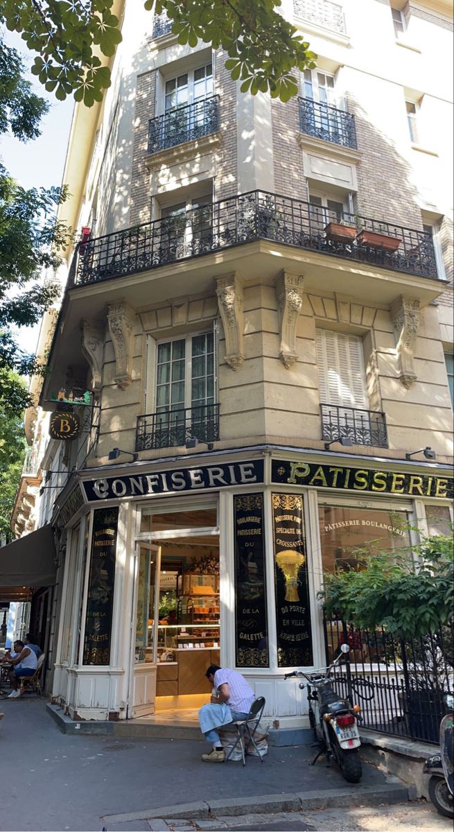 two people sitting on the sidewalk in front of a building with balconies and windows