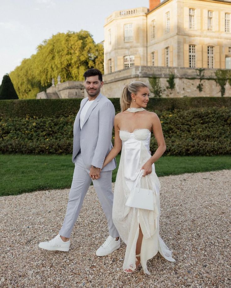 a man and woman dressed in formal wear walking down a gravel road near a large building