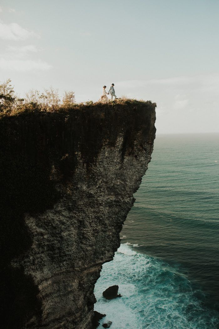 two people are standing on the edge of a cliff overlooking the ocean and cliffs below