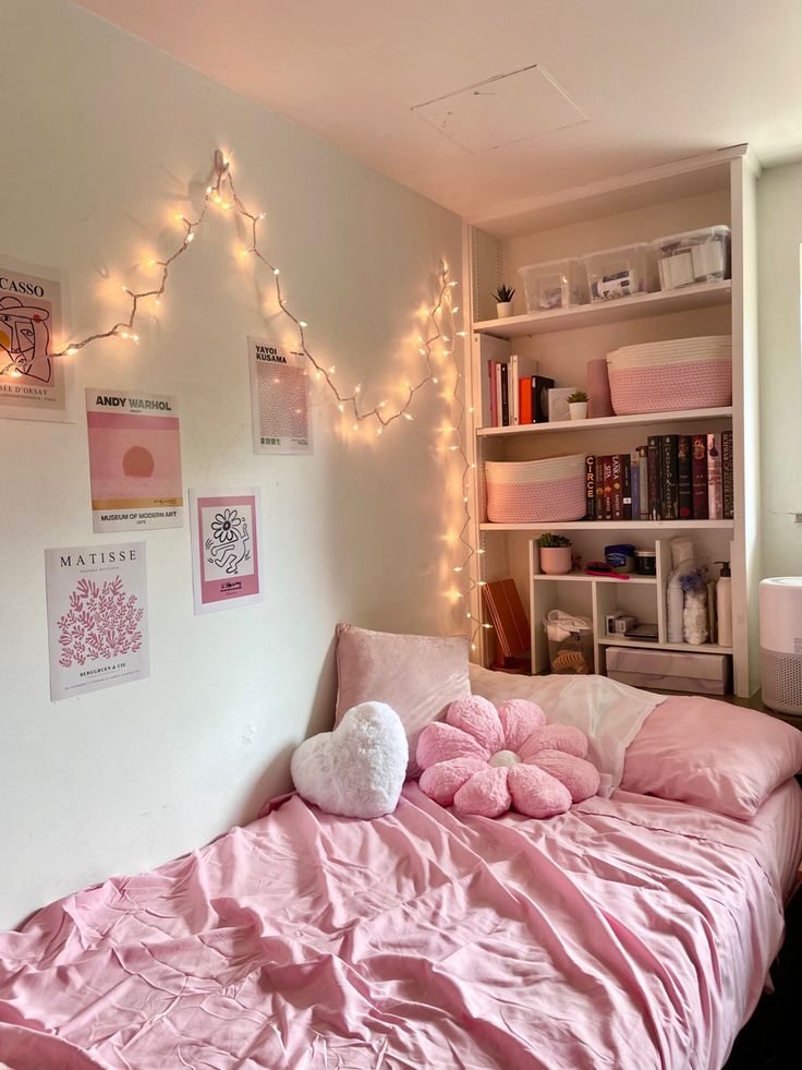 a bed with pink sheets and pillows in a room decorated with lights, bookshelves and pictures on the wall