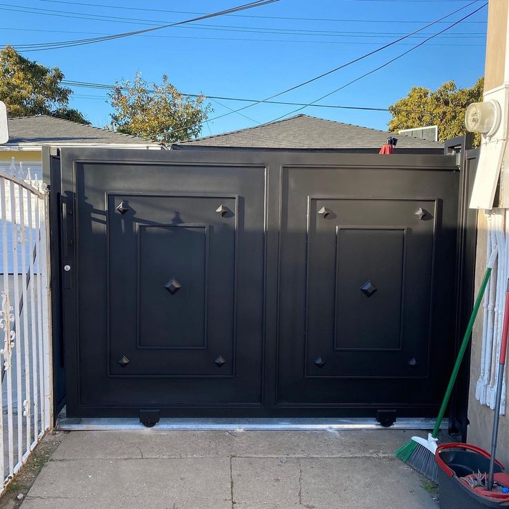 a black gate is open in front of a white fence and some trash cans on the sidewalk