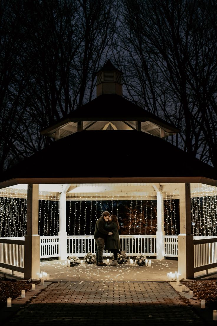 two people sitting on a bench in front of a gazebo with string lights at night