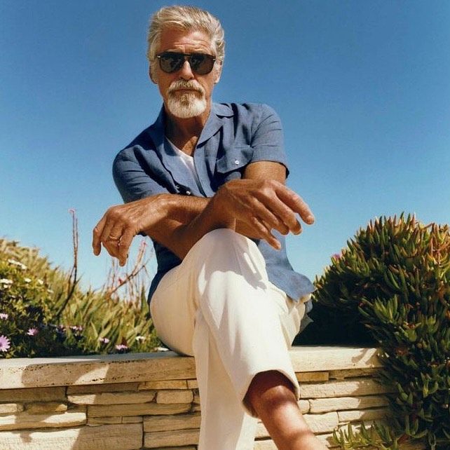 an older man sitting on top of a stone wall next to bushes and flowers with his legs crossed