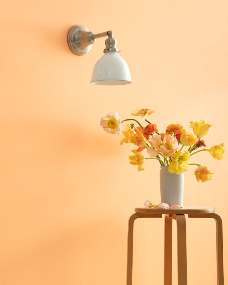 a white vase with yellow and red flowers on a table next to a wall light