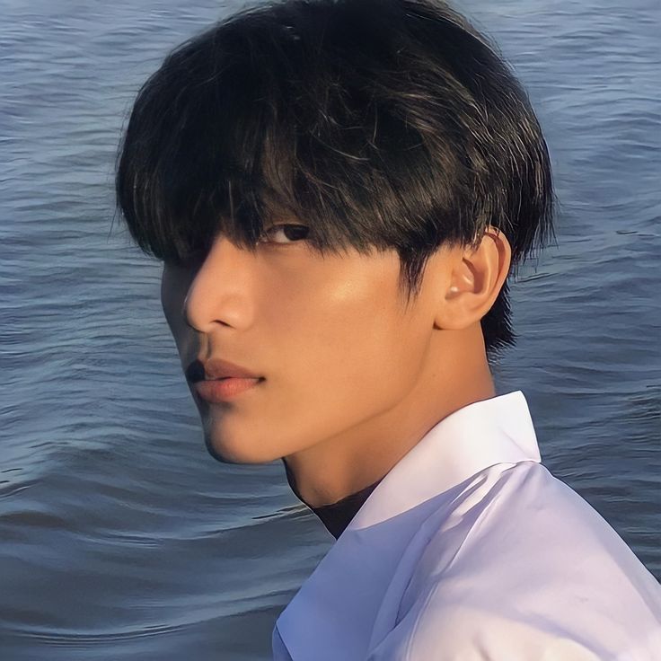 a man with black hair standing in the water looking off into the distance while wearing a white shirt