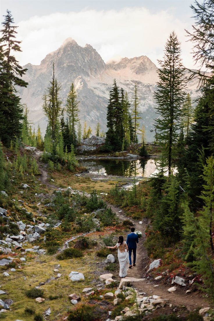 a man and woman walking down a trail in the woods with mountains in the background