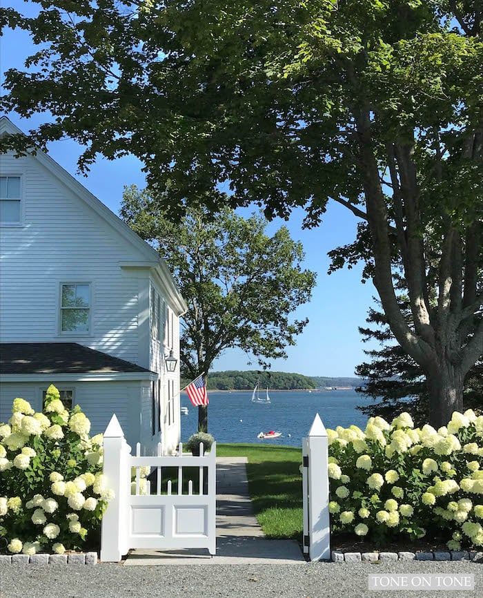 a white picket fence in front of a house with flowers and trees around it on a sunny day