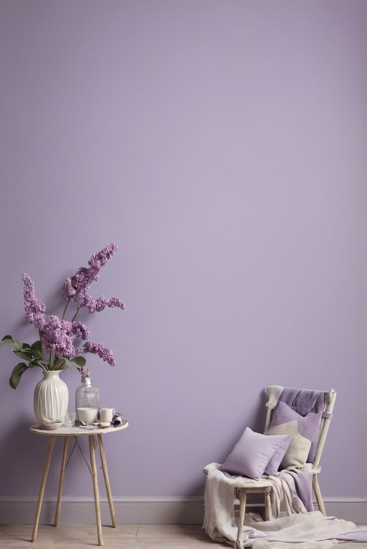 a chair and table with flowers on it in front of a purple wall, next to a white vase