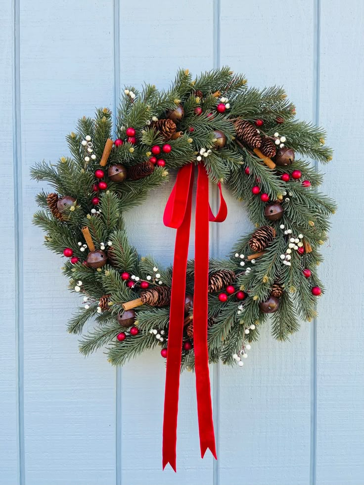 a christmas wreath hanging on the side of a blue door with red ribbon and pine cones