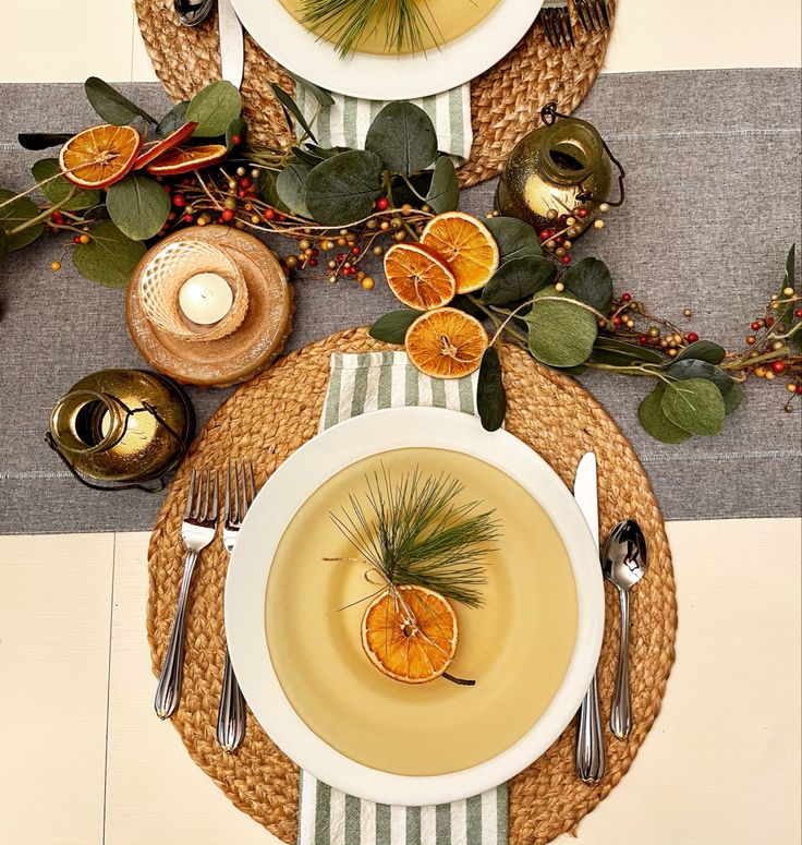 a place setting with oranges and silverware on a striped table cloth, surrounded by greenery