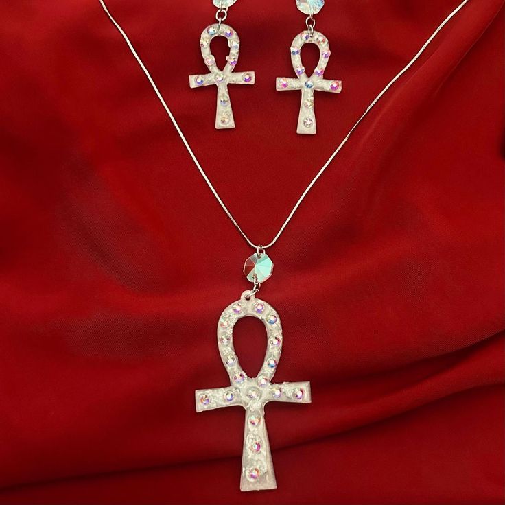 Our Ankh earring and necklace set is a stunning fusion of timeless symbolism and modern craftsmanship, meticulously crafted to embody the essence of elegance and mystique. At the heart of each piece lies the iconic Ankh symbol, revered in ancient Egyptian culture as a symbol of life and eternity. Sculpted with precision from luminous silver white mica, every Ankh pendant emanates a celestial glow, reminiscent of moonlit whispers on a tranquil night. Enveloped in hand-poured resin, the Ankh is lovingly encased, preserving its significance and beauty for generations to come. Delicate veins of silver leaf delicately adorn the edges, enhancing the ethereal allure of the design, while tiny rhinestones add a touch of celestial sparkle, reminiscent of stars scattered across a velvet sky. The earr Silver Crystal Spiritual Jewelry, Spiritual Crystal Jewelry Gift, Spiritual Crystal Jewelry For Gifts, Spiritual Crystal Jewelry As A Gift, Nickel-free Ankh Elegant Jewelry, Nickel-free Ankh Shaped Elegant Jewelry, Nickel Free Ankh Jewelry Gift, Elegant Nickel-free Ankh Jewelry, Nickel-free Ankh Jewelry