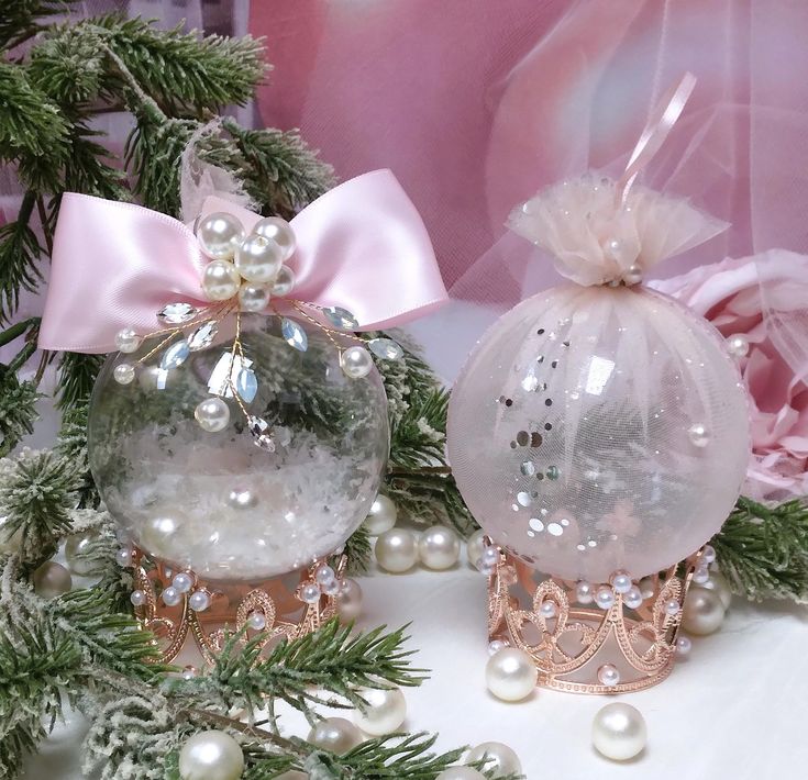 two glass ornaments with pink bows and pearls on them sitting next to some pine branches