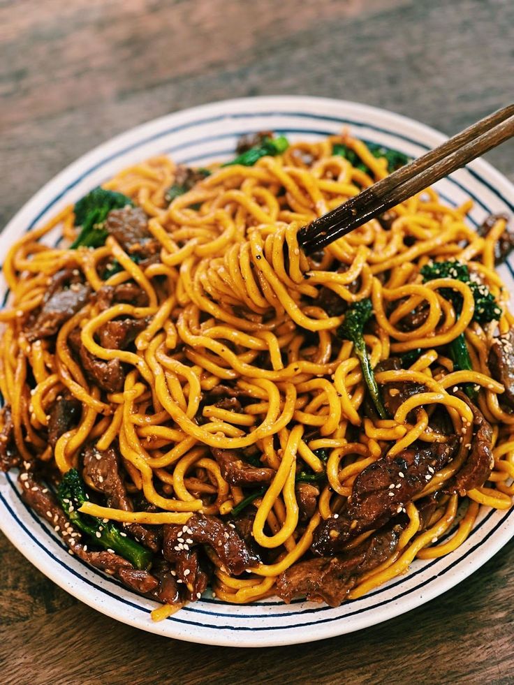 a white plate topped with noodles and beef next to chopsticks on top of a wooden table