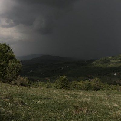 a dark cloud hovers in the sky over a grassy field with trees and mountains