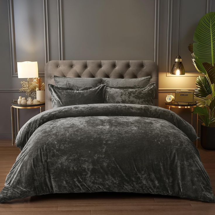 a bed with grey comforter and pillows in a room next to a lamp on a table