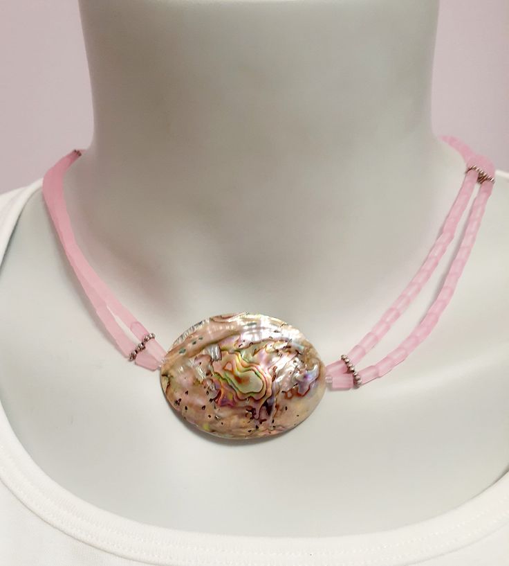 This abalone shell and pink bead short necklace is the perfect piece to add a touch of boho chic style to any outfit. The necklace features 2 strands of pink cylindrical beads, giving it a unique and eye-catching look. The abalone shell bring s a subtle iridescent shimmer to the necklace, while the pink beads compliment the dome shaped abalone shell.  Whether you're looking for a statement piece or something more subtle, this abalone shell and pink bead short necklace is the perfect addition to any jewelry collection. Adjustable Double Strand Pink Jewelry, Adjustable Round Beads Shell Necklace, Adjustable Pink Shell Jewelry, Pink Adjustable Shell Jewelry, Adjustable Multi-strand Pink Jewelry, Adjustable Pink Choker For The Beach, Pink Shell Necklace With Round Beads, Pink Shell Necklace With Round Beads As Gift, Pink Shell Necklace With Round Beads For Gift