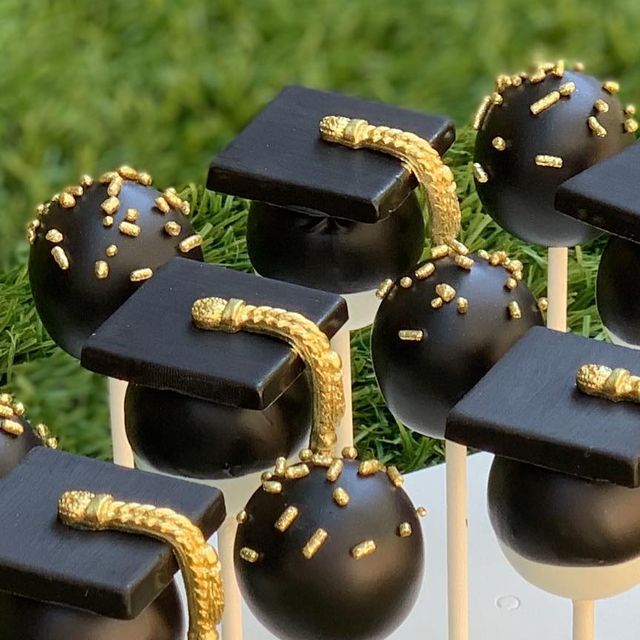 graduation cake pops decorated with black and gold graduation caps on top of white sticks in front of green grass