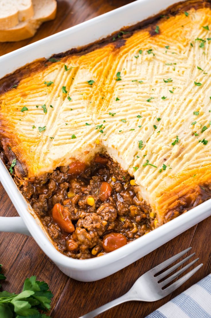 a casserole dish with meat and vegetables in it on a wooden table next to bread