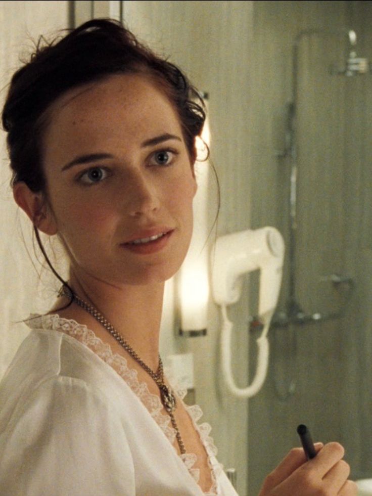 a woman in a white dress is holding a pen and looking at the camera while standing in front of a bathroom mirror