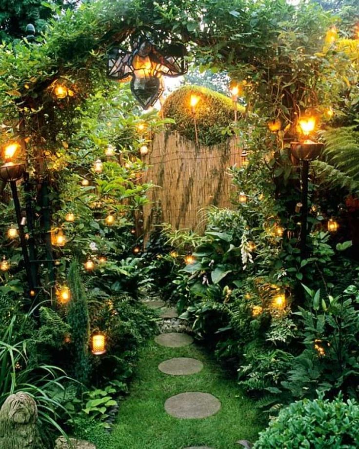 a garden with lots of green plants and lights on the trees, along with stepping stones