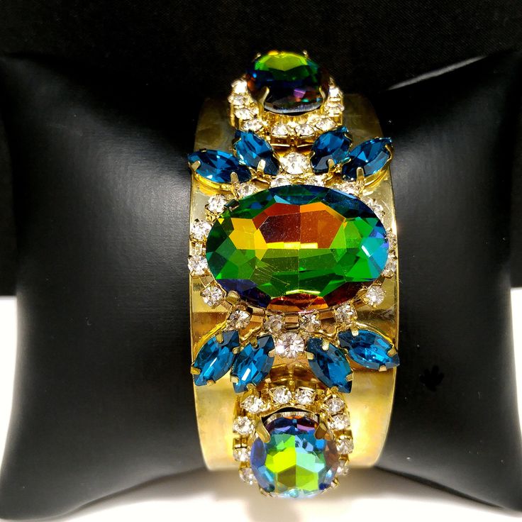 *Description: This is a gorgeous watermelon rhinestone crystal bracelet on a golden brass cuff which glitters and sparkles in the light. The vintage rhinestones are secured on a golden brass cuff and the reflective cuff adds additional sparkle to the bracelet. There is one large watermelon rhinestone in the center, surrounded by clear rhinestones and sapphire blue marquise cut rhinestones. On each side of the center stone are two more watermelon rhinestones, a little smaller in size, one on each Metal Crystal Bracelet With Stones For Party, Party Crystal Bracelet With Metal Stones, Crystal Bangle Cuff Bracelet For Party, Party Crystal Bracelet With Stones, Glamorous Crystal Cuff Bracelet For Party, Crystal Bracelet With Jewels For Parties, Party Cuff Bracelet With Crystal Bling, Crystal Bracelets With Jewels For Parties, Green Stone Bracelets For Party