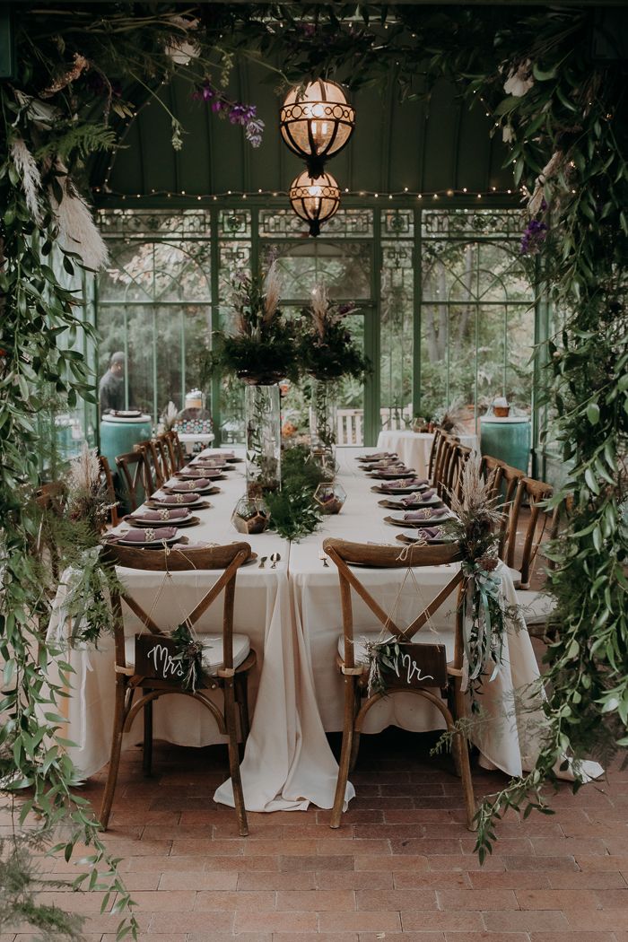 a table set up for a formal dinner with greenery on the walls and chairs