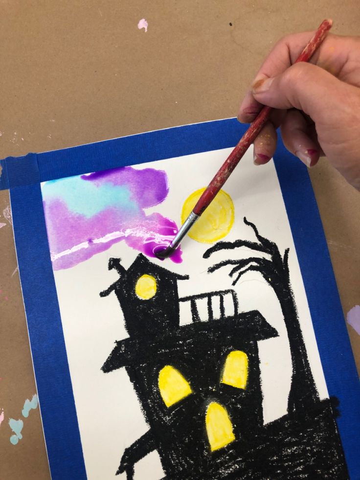 a child's hand holding a paintbrush over a drawing of a house on paper