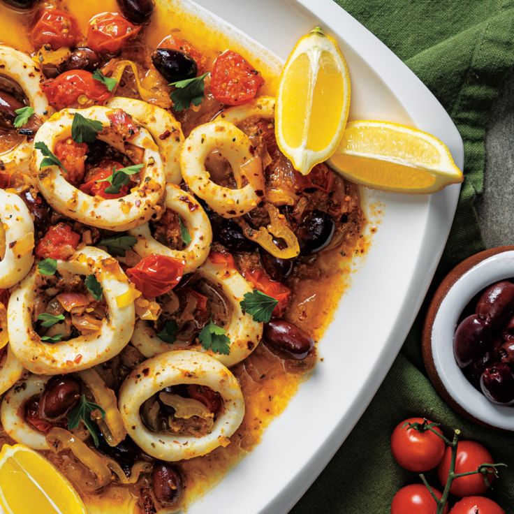 a white plate topped with cooked squid and olives next to tomatoes, lemon wedges and cherry tomatoes