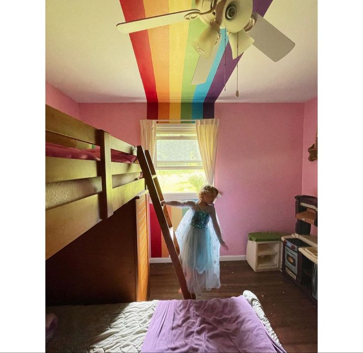 a girl in a blue dress is standing on a bed with a rainbow painted ceiling fan