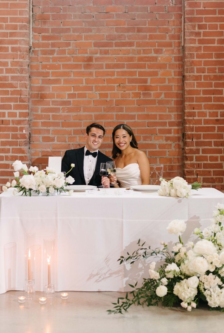 a bride and groom sitting at a table in front of a brick wall with candles